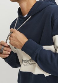 Hoodie bicolore bleu marine et crème avec une poche frontale, capuche à cordon avec cordon décoré, poignets côtelés et détail de logo imprimé sur la poitrine.