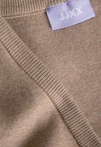 Beige gebreide trui met geribbelde rand en een lichtpaarse kledinglabel met de witte tekst "JJXX".