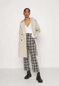 Manto bege sobre uma blusa branca envolvente, combinado com calças xadrez preto e branco e botas de plataforma pretas. Teclado suave e design estruturado.