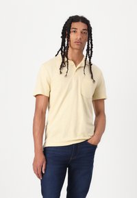 Polo skjorte i lys gul bomull med kortermet, spiss krage, enkelt brystlomme og teksturert stoff. Kombinert med mørke jeans.