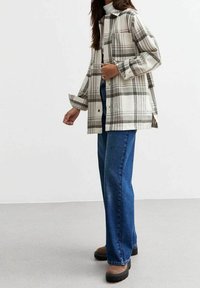 Giacca a camicia a quadri nei toni crema, grigio e nero. Presenta un colletto, chiusura con bottoni e spacchi laterali. Abbinata a jeans larghi blu e stivali marroni.
