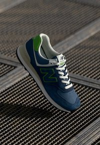 Navy suede sneaker with green accents, white laces, and logo. Features a cushioned sole and a textured side design. Model 574.