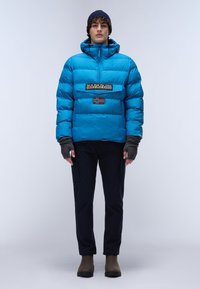 Dikke blauwe jas met logo, voorzien van een rits aan de voorkant, verstelbare capuchon en grijze vingerloze handschoenen; gecombineerd met donkere broek en bruine laarzen.