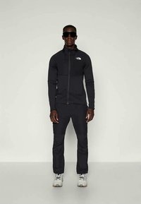 Casaco atlético preto com fecho, combinado com calças a condizer. Logótipo refletor no casaco; o modelo usa óculos de sol e sapatos claros.