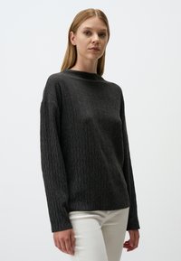 Pull en tricot gris avec un col montant, présentant un motif en diamant texturé et des manches longues, associé à un pantalon de couleur claire.