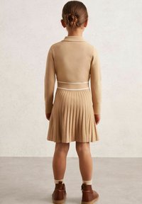 Beež plisseeritud kleit sobiva peal-img, valge vöökoht ja pikad varrukad. Kantud pruunide pahkluu saapade ja valgete sokkidega.