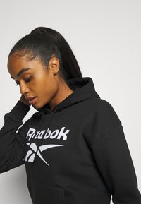 Czarna hoodie z kapturem wykonana z mieszanki bawełny. Posiada duże białe logo Reebok z przodu, kieszeń typu kangur, oraz ściśnięte mankiety.