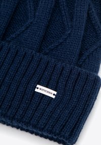 Námořnicky modrá pletená čepice s žebrovaným vzorem a vystouplým trojúhelníkovým motivem. Obsahuje stříbrnou cedulku s nápisem "WITTCEN".