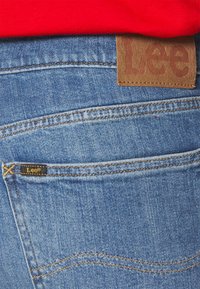 Vaqueros de mezclilla azul con un parche de cuero marrón en la parte trasera de la cintura marcado "Lee". Presenta costuras doradas y una pequeña etiqueta negra en el bolsillo trasero.