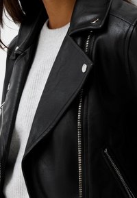 Czarna skórzana kurtka motocyklowa z metalowymi zamkami błyskawicznymi i zatrzaskami, o textured wykończeniu i szerokimi klapami. Nosi się ją na jasnoszarym swetrze.