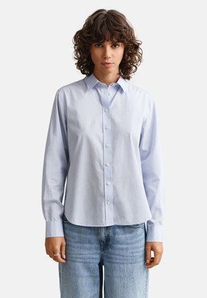 Γυναίκα με σγουρά σκούρα μαλλιά που φοράει ένα ανοιχτό μπλε πουκάμισο με κουμπιά και ανοιχτό μπλε τζιν, στέκεται μπροστά σε ένα απλό ανοιχτό φόντο.