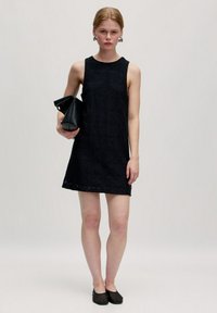 Schwarzes ärmelloses Kleid mit strukturiertem Muster, knielang, kombiniert mit einer schwarzen Clutch und flachen schwarzen Schuhen.