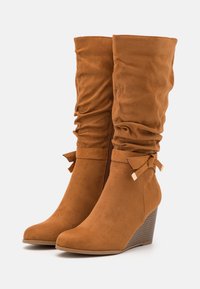 Stivali a zeppa in suede marrone con design sbiadito, caratterizzati da un fiocco decorativo e un tacco zeppa texturizzato.