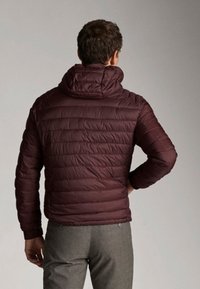 Burgundy pufferjakke med hætte, der har horisontale quiltede detaljer og en tætsiddende design, parret med grå bukser.