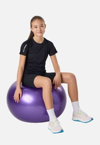 Jeune fille en tenue de sport noire assise sur un ballon d’exercice violet, portant des chaussettes blanches et des chaussures de sport multicolores, sur fond blanc.