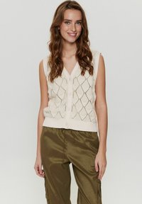 Stickad ärmlös väst i krämfärg med ett mönster av diamantöppningar och knappstängning framtill. Matchas med olivgrön, blank bred byxa.