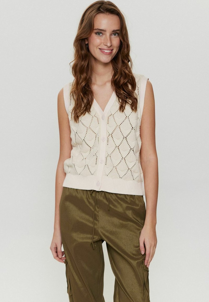 Stickad ärmlös väst i krämfärg med ett mönster av diamantöppningar och knappstängning framtill. Matchas med olivgrön, blank bred byxa.