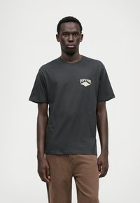 Melna kokvilnas T-kreklā ir neliels balts un zaļš Rip Curl logo kreisajā krūtī. Ar īsām piedurknēm un klasisku apaļu kakla dizainu.