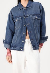 Jeansowa kurtka w głębokim niebieskim kolorze z klasycznym kołnierzykiem, zapięciem na guziki, dwoma przednimi kieszeniami i podwiniętymi rękawami, noszona na czerwoną koszulę w kratkę.