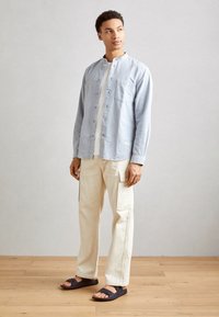 Svijetloplena košulja na dugih rukava s okovratnikom, bež cargo hlače i crne sandale. Glatka tekstura tkanine i opušteni kroj.