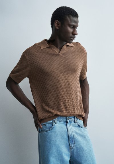 Homme en polo marron à manches courtes texturé et jean bleu clair, debout les mains derrière le dos, regardant sur le côté sur un fond gris uni.