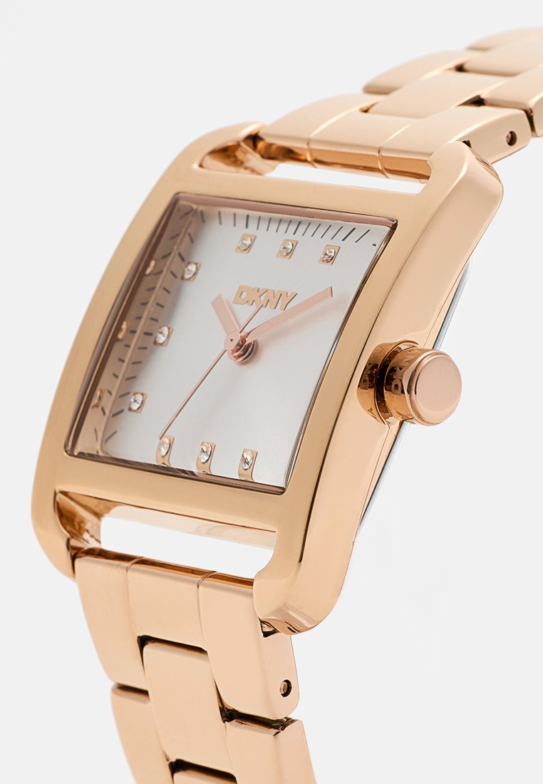 Reloj rectangular DKNY en oro rosa con esfera blanca, marcadores de cristal y una pulsera de metal pulido con cierre de broche.