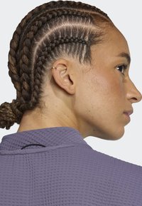 Prenda tejida de color púrpura con un patrón texturizado, con un cuello alto y un pequeño acento negro en la parte posterior. El cabello está peinado en trenzas.