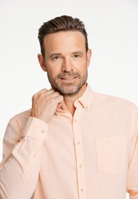 Camisa de botones en melocotón claro con una tela texturizada, que presenta un bolsillo en el pecho y botones en la parte delantera. El modelo toca suavemente el cuello.