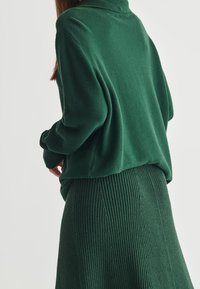 Zielony sweter robiony na drutach z wysokim kołnierzem i luźnym krojem, zestawiony z prążkowaną spódnicą ołówkową, podkreślającą teksturowany materiał.