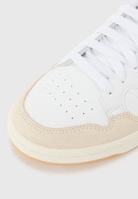 Zapatilla blanca con un upper de cuero liso y acentos de ante beige. Punta redonda, diseño perforado y cordones blancos con una suela clara.