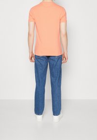 T-shirt a maniche corte color pesco abbinato a jeans a gamba dritta blu e sneakers bianche, visto da dietro su uno sfondo chiaro.