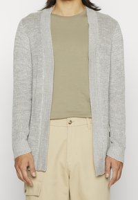 Hellgraue Strickjacke mit strukturiertem Strickmuster, offenem Vorderteil und gerippten Bündchen, getragen über einem beigen Shirt und hellbeigen Hosen.