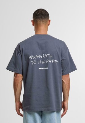 Dark gray cotton t-shirt with short sleeves, featuring white text on the back that reads, "ALWAYS LATE TO THE PARTY." Casual fit.