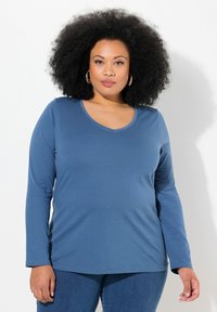 Dlouhý rukáv modrého trička s výstřihem do V, vyrobeného z měkké tkaniny. Má uvolněný střih a ploché švy. V kombinaci s modrými džíny.