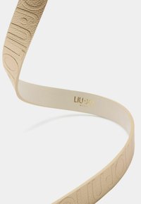 Beżowy skórzany pasek z wytłoczonym logo, charakteryzujący się gładką teksturą i błyszczącym złotym akcentem nazwy marki na wewnętrznej stronie.