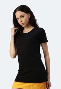 Schwarzes Kurzarm-T-Shirt aus glattem Stoff mit Rundhalsausschnitt, kombiniert mit einem plissierten gelben Rock, vor einem hellen Hintergrund.