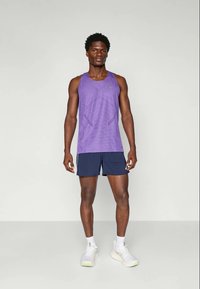 Lila ärmelloses Sporttanktop mit geometrischem Muster, kombiniert mit marineblauen Shorts. Das Outfit besteht aus weißen Sneakers mit grünen Akzenten.