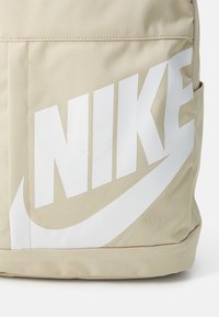 Beige stoffen rugzak met groot wit Nike-logo. Heeft een gestructureerd oppervlak en versterkte stiksels langs de randen en zakken.