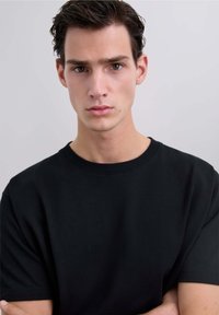 Tricou din bumbac negru cu decolteu rotund și mâneci scurte, având o textură fină și o croială lejeră, purtat de un model.