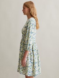 Robe fleurie avec un fond jaune clair et des motifs de fleurs bleues et vertes. Elle a des manches trois-quarts et une taille froncée.