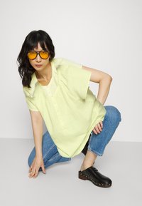 Blusa amarela clara de mangas curtas com fecho de botões e tecido texturizado, combinada com jeans de ganga azuis e clogs pretos com detalhes em metal.