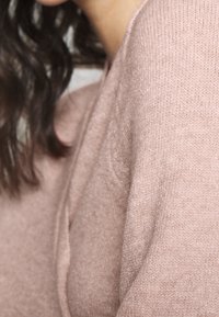 Světle růžový pletený svetr s jemnou texturou; má jemný lesk a uvolněný střih, který zvýrazňuje detailní vzor látky.