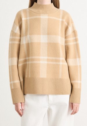 Pull en maille à carreaux beige et blanc avec col haut, poignets et ourlet côtelés, porté avec un pantalon blanc, visible des épaules aux hanches.