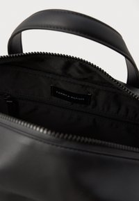 Zwarte lederen handtas met een ritssluiting, binnenzakken en een label met de tekst "TOMMY HILFIGER." Glad oppervlak en stevige constructie.