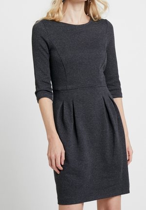 Femme portant une robe ajustée, gris foncé, texturée, à manches trois-quarts avec des plis à la taille, debout devant un fond clair uni.