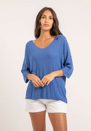 Femme aux longs cheveux bruns portant un pull bleu côtelé et un short blanc retroussé, debout devant un fond clair uni.