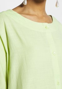 Donna che indossa una camicia verde pallido con bottoni e scollatura rotonda, con grandi orecchini di perle, ripresa dalla parte del collo fino al petto.