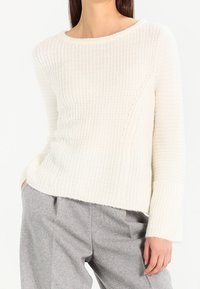 Valkoinen neulottu neule, jossa on ribbineulos, pyöreä pääntie ja pitkät hihat, yhdistetty harmaisiin, väljiin housuihin.