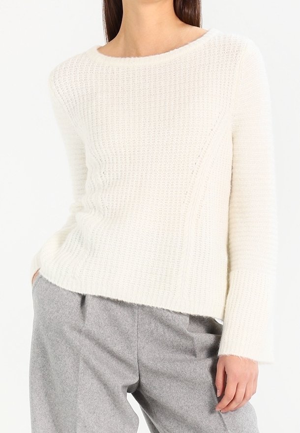 Bílý pletený svetr s žebrovanou strukturou, kulatým výstřihem a dlouhými rukávy, v kombinaci se šedými, volně střiženými kalhotami.