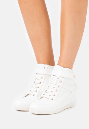 Białe sneakersy sięgające do kostki z obcasem na koturnie, z złotymi elementami, sznurowanym przodem, paskiem na rzep oraz teksturowanymi akcentami po bokach.